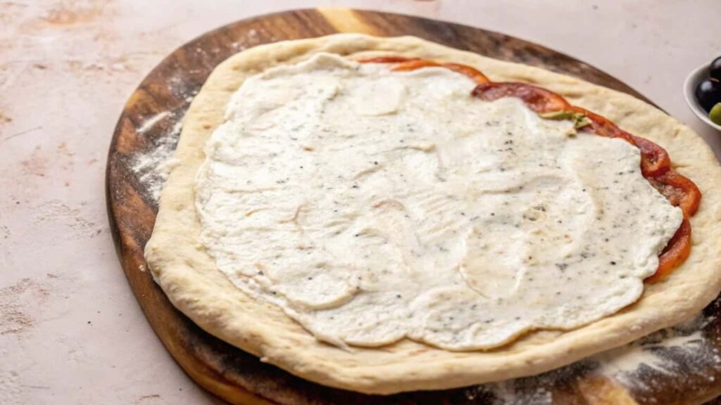 Unbaked pizza dough on a wooden board, topped with creamy garlic sauce and layered with sliced tomatoes.

