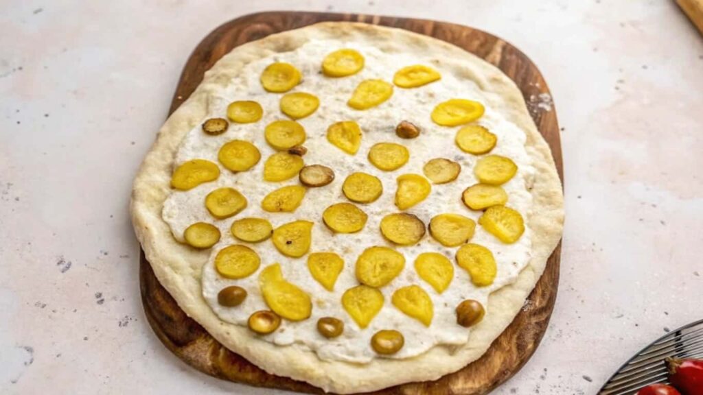 Unbaked pizza on a wooden board, topped with creamy garlic sauce and evenly spread sliced pickles.