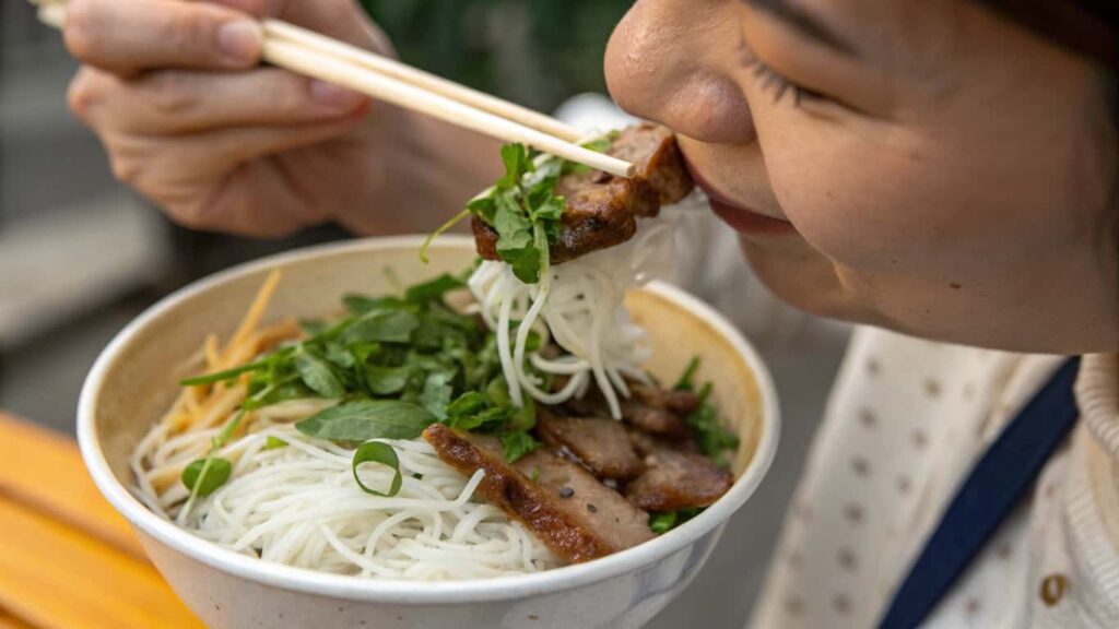 A woman enjoying a bowl of Vietnamese noodle dish with fresh herbs and grilled meat.